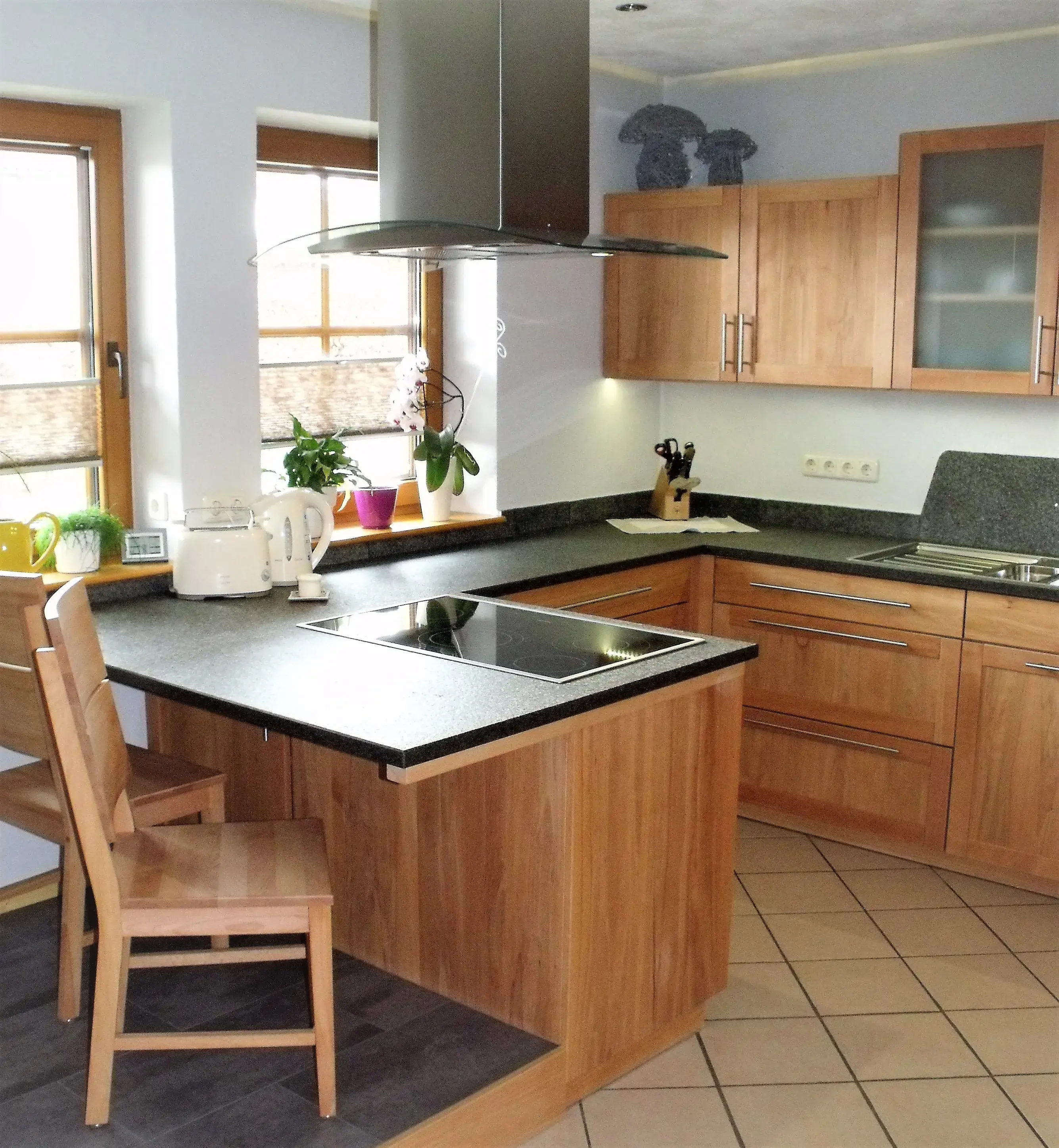 Helle Einbauküche mit Holzschränken, Kochinsel, Stuhl und großen Fenstern.