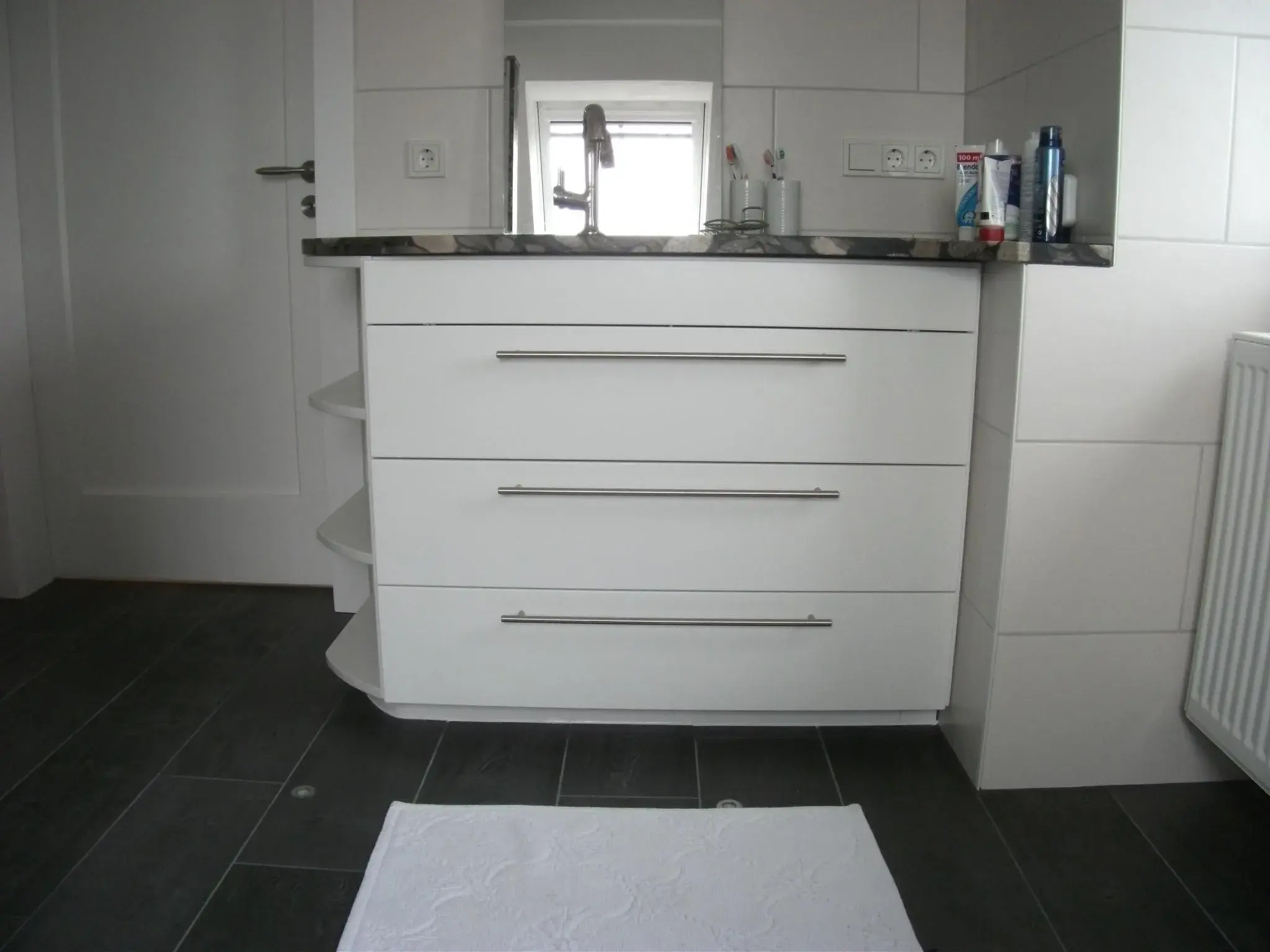 Weißer Badunterschrank mit drei Schubladen, dunkle Arbeitsplatte, Spiegel darüber.