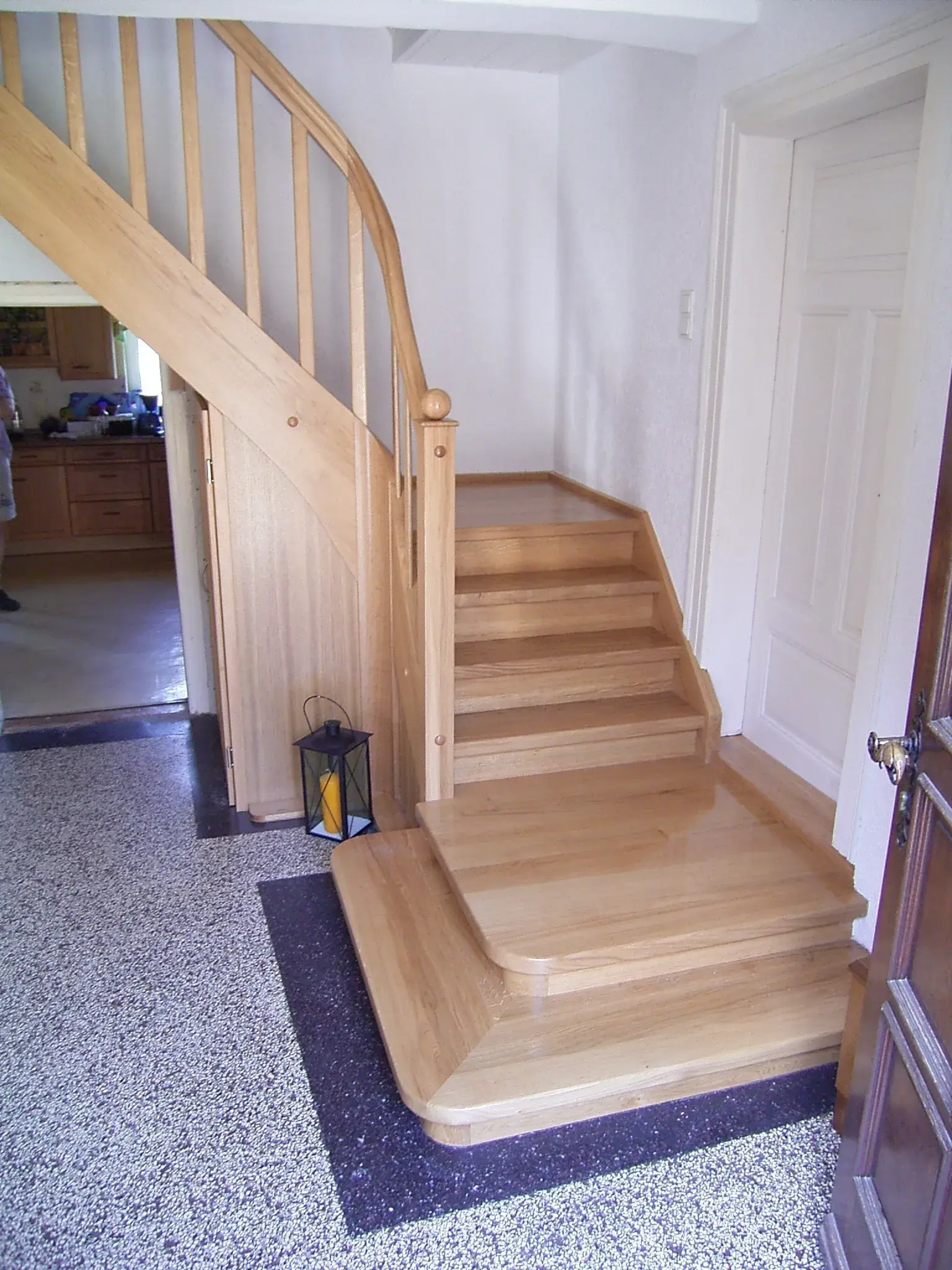 Holztreppe im Flur mit Geländer, daneben weiße Tür und gemusterter Boden.