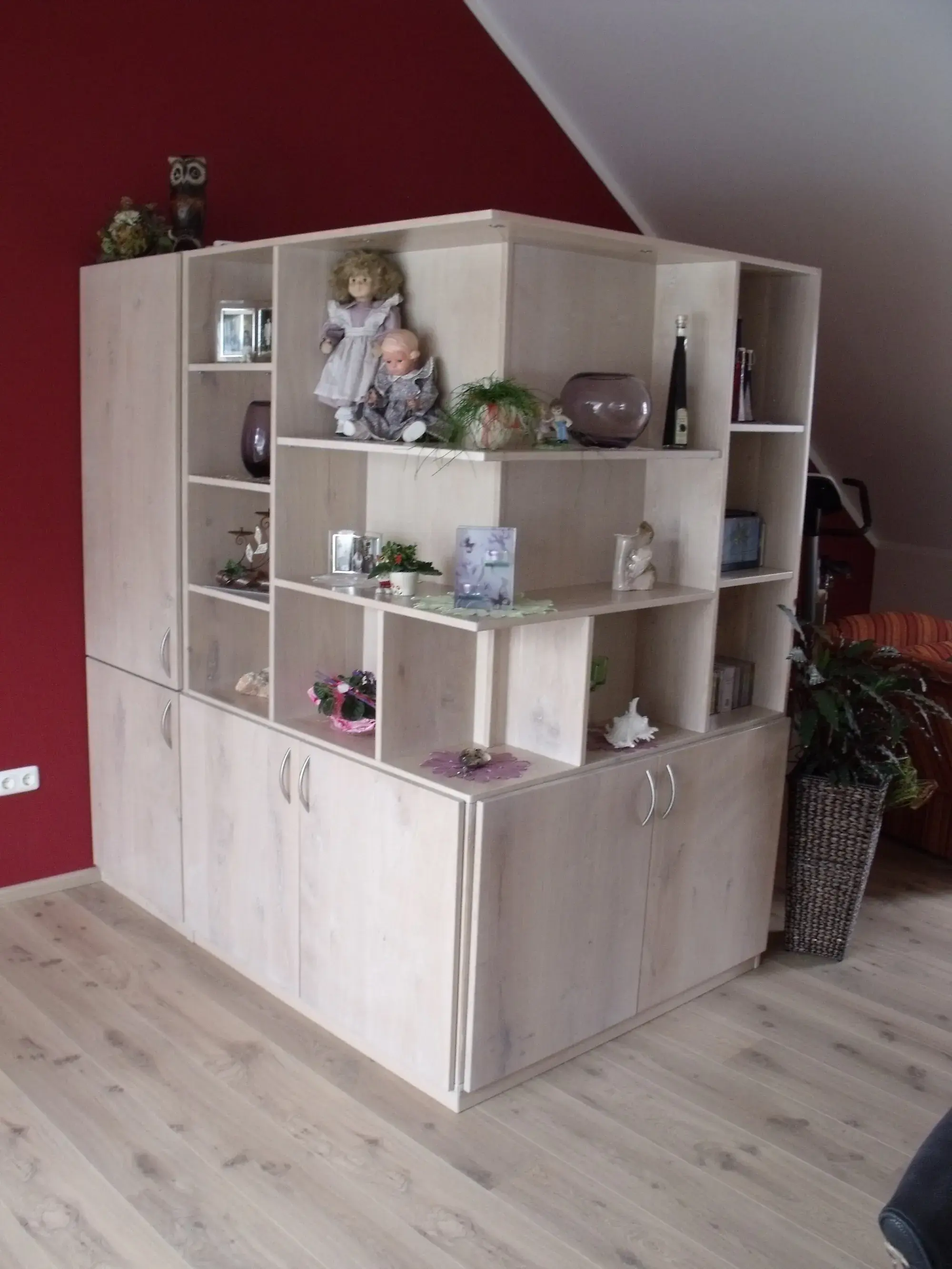 Holzregal mit offenen Fächern, dekoriert mit Figuren, Pflanzen und Karten vor roter Wand.