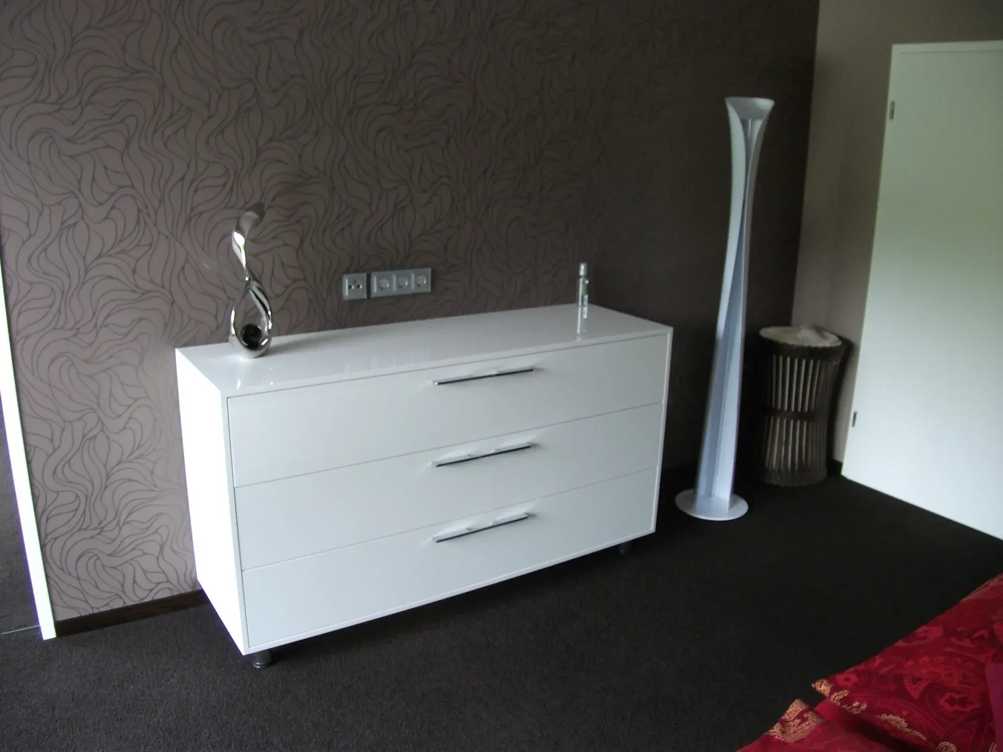 Weiße Kommode mit Dekoration, moderne Stehlampe daneben, dunkler Teppichboden.