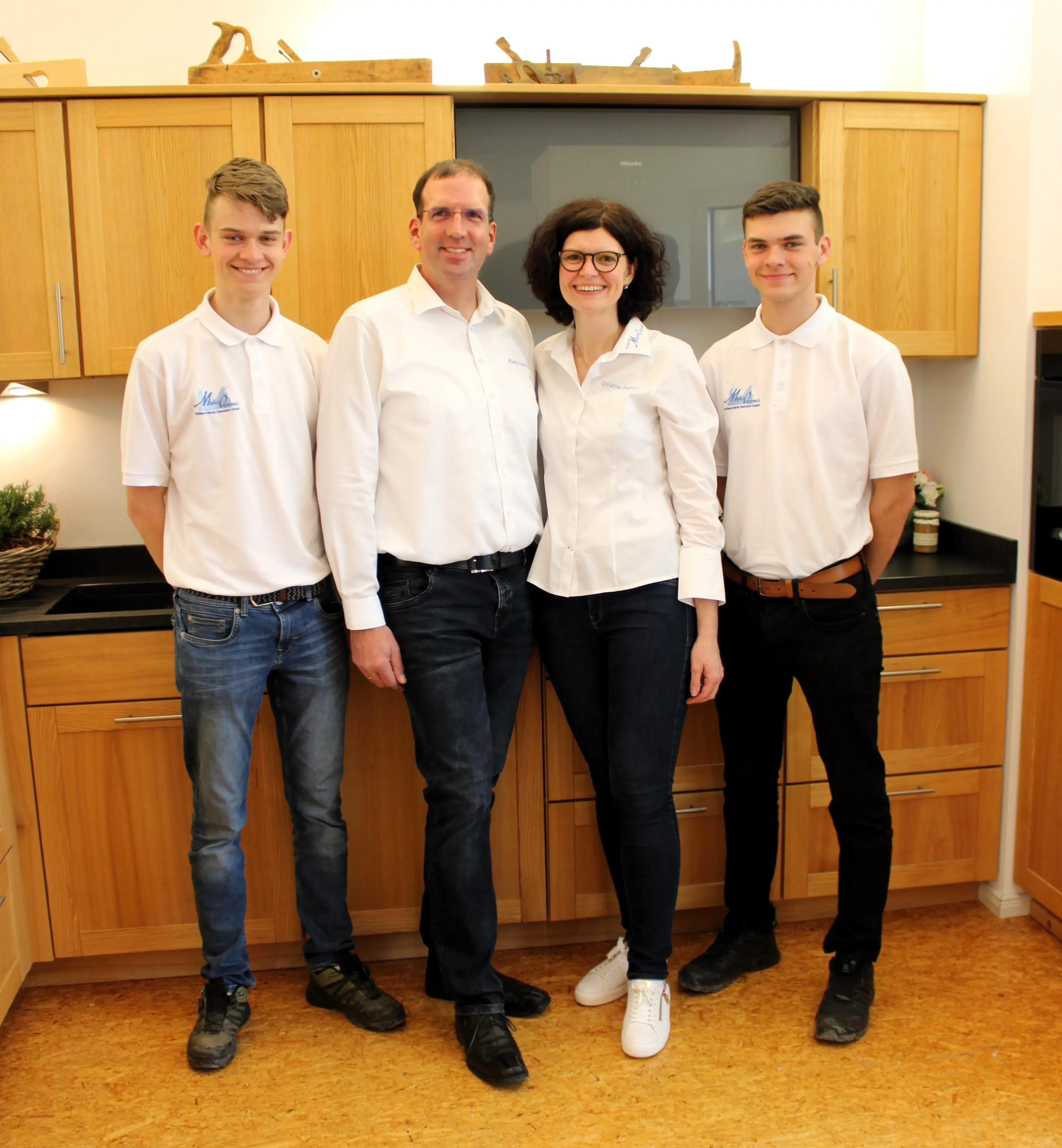 Vier Personen in weißer Kleidung stehen lächelnd in einer modernen Holzküche.