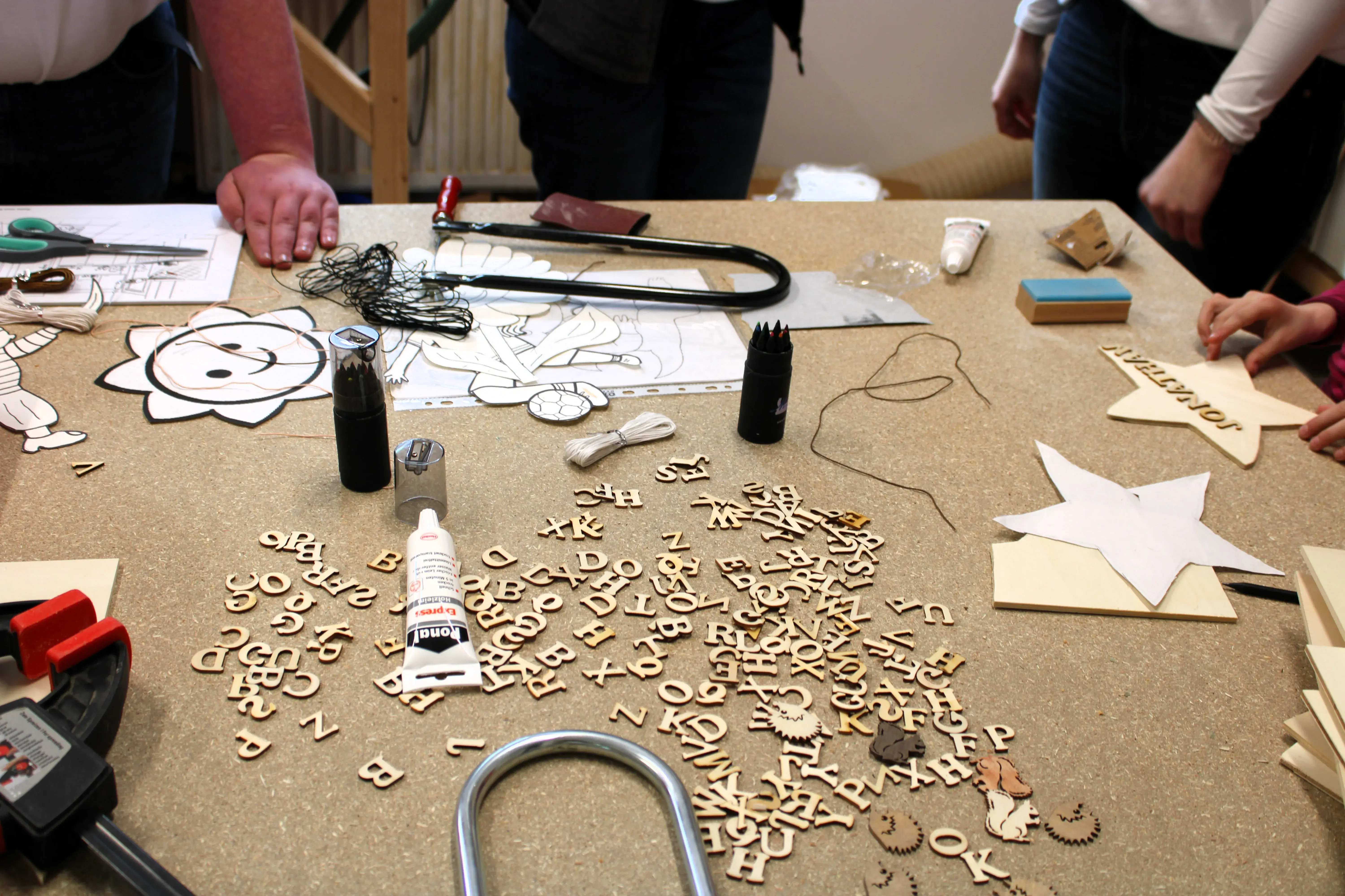 Basteltisch mit Holzbuchstaben, Werkzeug, Sternen und mehreren Händen.