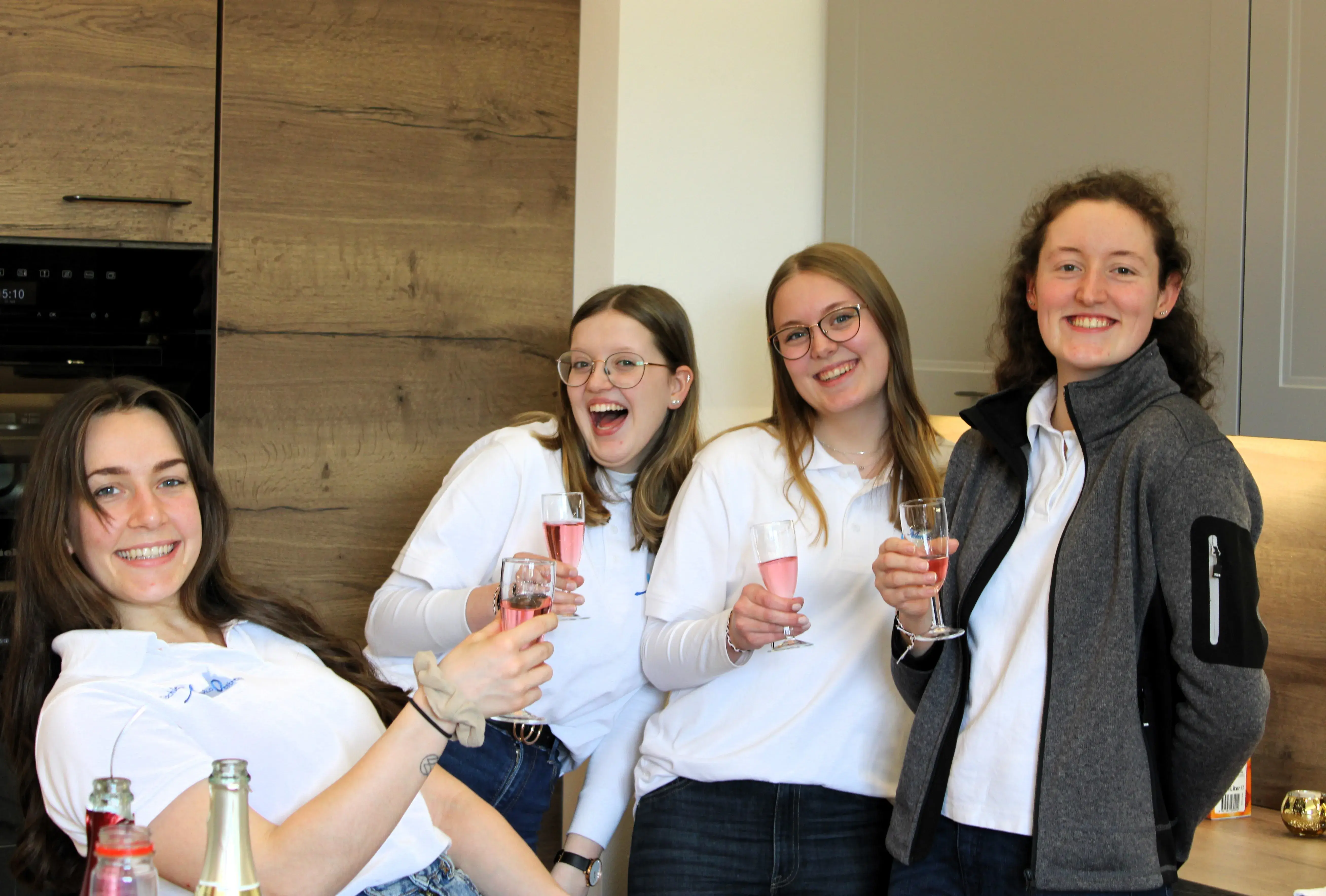 Vier junge Frauen lächeln in einer Küche und stoßen mit Getränken an.