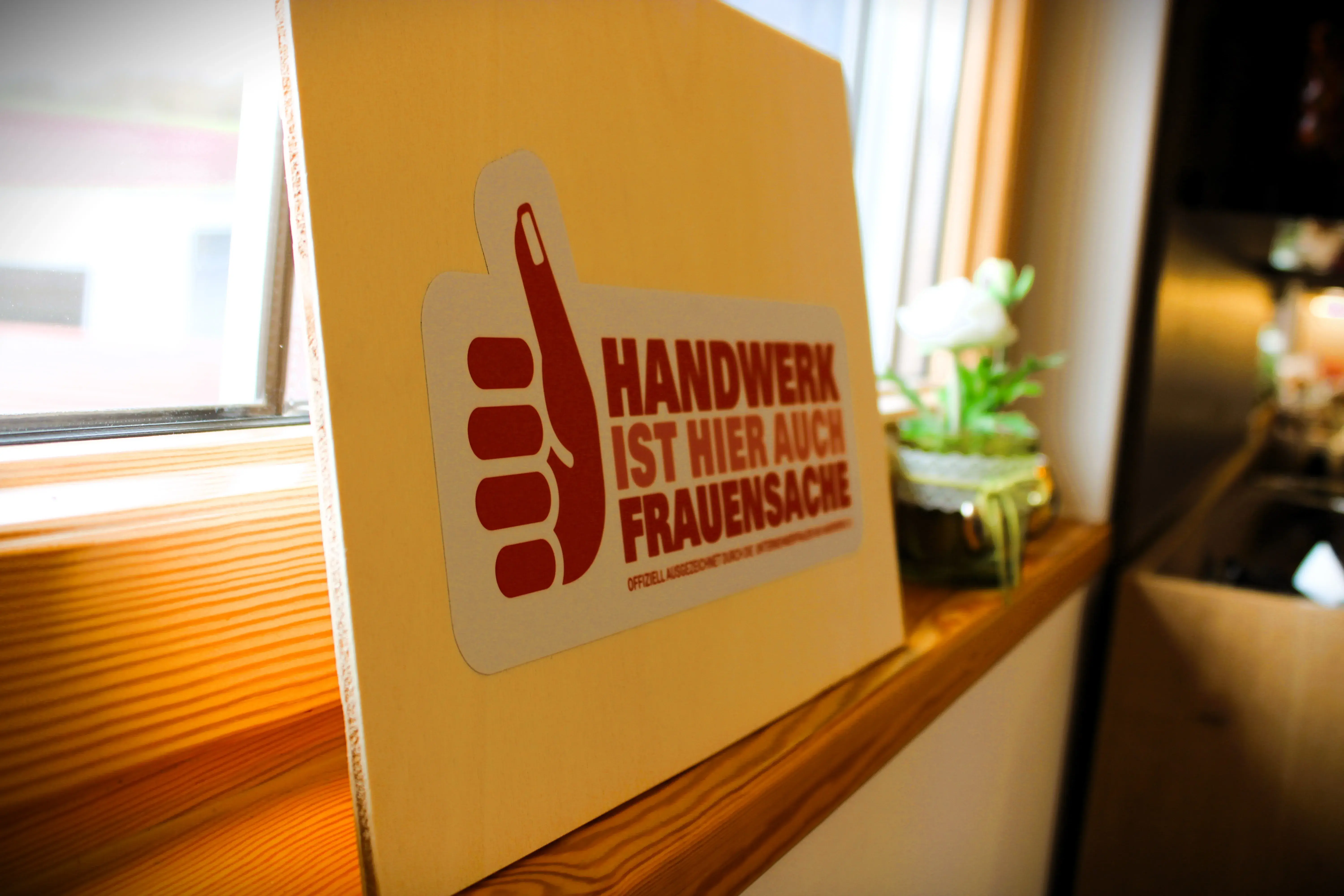 Schild mit rotem Daumen und Text „Handwerk ist (k)ein Frauensache“ auf Fensterbank.